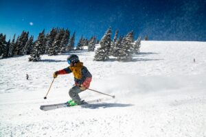 Expert skier carving down a snow-covered ski slope under a bright blue sky with snow-covered pine trees in the background.